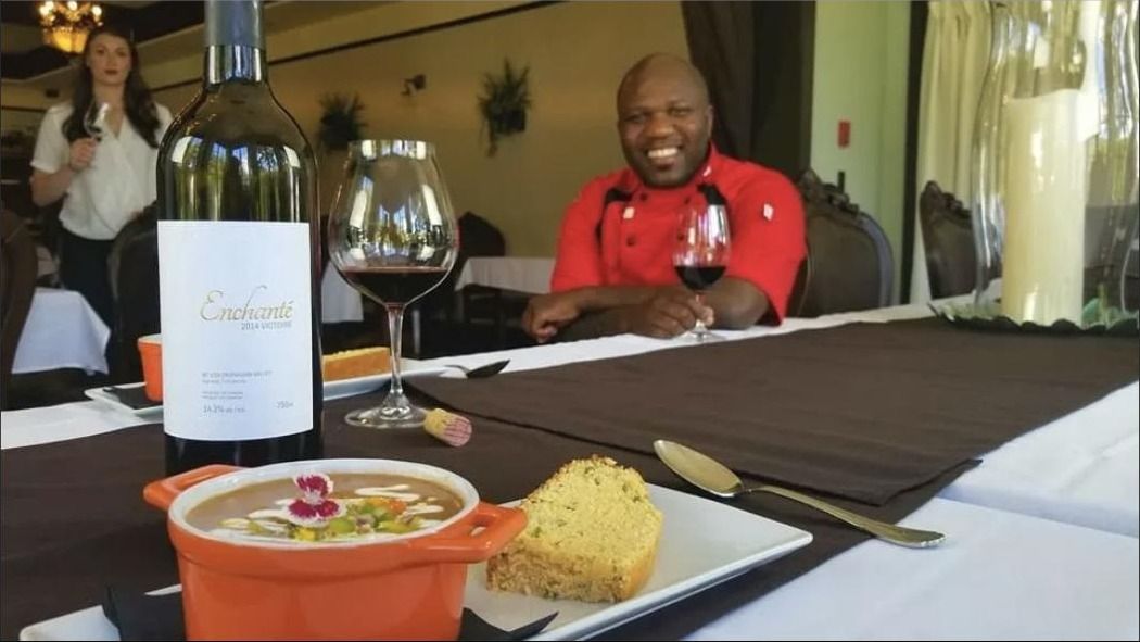 A chef seated behind a plated dish and a bottle of wine during a promotional shoot